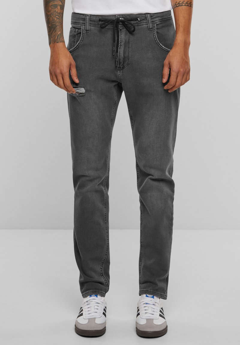 Szürke farmernadrág, slim fit szabással, koptatott térddel, zsinóros derékrésszel és eltérő varrási részletekkel. Fehér sportcipővel viselve.
