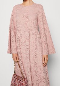 Sukienka z koronki w jasnym różu z długimi rękawami, kwiatowymi wzorami i dopasowanym górnym częścią, w połączeniu z wielokolorową torbą plecioną z łańcuszkiem.