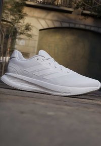 Weißer athletischer Laufschuh mit drei Seitenstreifen und Cloudfoam-Sohle, platziert auf einer Außenpflasterfläche.