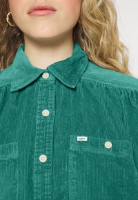 Femme portant une chemise verte en velours côtelé, boutonnée, avec un col et une petite étiquette de la marque Lee sur la poche poitrine gauche.