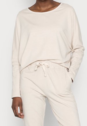 Personne portant un haut beige ample à manches longues et un pantalon beige assorti avec un cordon, debout avec une main dans la poche.