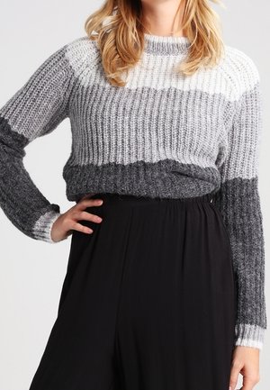Femme portant un pull en maille dégradé gris avec texture côtelée et pantalon noir ample taille haute, main reposant sur la hanche.
