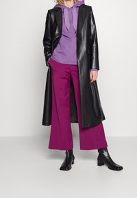 Abrigo de cuero negro sobre una sudadera morada y pantalones morados de pierna ancha. Botines negros, diseño estilizado y texturas suaves. Fondo neutro.