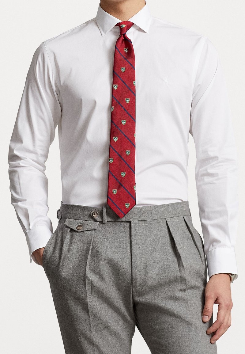 Camicia bianca con colletto button-down, abbinata a una cravatta rossa con accenti verdi e blu, e pantaloni grigi a pieghe con doppio bottone.