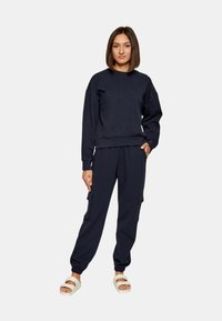 Felpa blu navy e pantaloni jogger coordinati, con tasche cargo, polsini a coste e un motivo testurizzato con logo. Abbinati a scarpe slip-on bianche.