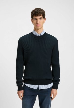 Dark green ribbed sweater with a crew neck, paired with a light blue striped shirt underneath and dark denim jeans. Simple, textured design.