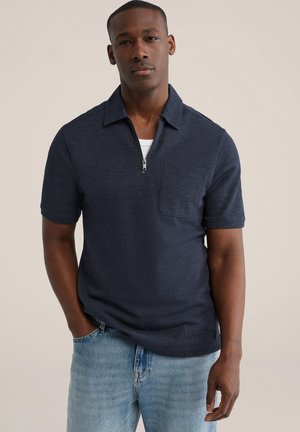 Man draagt een marineblauw poloshirt met korte mouwen, een rits aan de voorkant en een borstzak, gecombineerd met lichtblauwe spijkerbroek, staat met één hand in zijn zak.