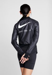 Veste légère noire avec des manches longues, des poignets élastiques et une texture brillante ; présente un logo Nike blanc et le texte "JUST DO IT" au dos.