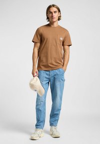 Brun kortärmad t-shirt med ficka, randiga blå cargobyxor och en beige keps. Vit logotyp-patch på t-shirten, sneakers på fötterna.