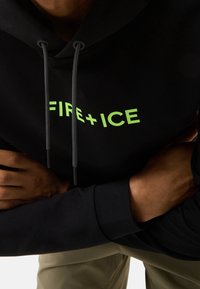 Personne portant un sweat à capuche noir avec le texte en vert néon « FIRE + ICE », bras croisés et pantalon beige visible sous la taille.