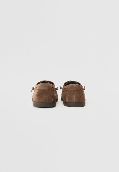 Brown suede loafers with a rounded toe and low rubber sole. Features decorative laces and stitched detailing at the heel.