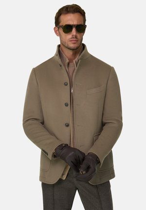 Homme portant des lunettes de soleil marron, un manteau beige boutonné sur un pull zippé, des gants foncés et un pantalon gris sur un fond uni.