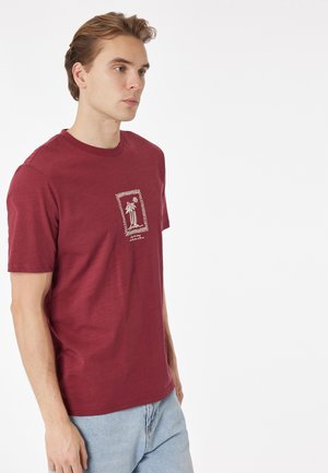 Giovane uomo che indossa una maglietta color granata con una grafica di palma e sole sul petto, abbinata a jeans azzurri, in piedi su sfondo bianco.