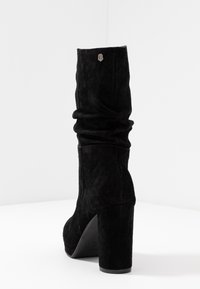 Black suede ankle boot with a slouchy design, thick high heel, and a silver logo accent on the back. Smooth texture with a matte finish.