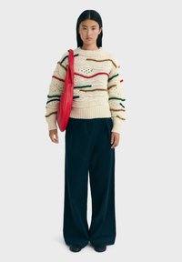 Cremefarbener Strickpullover mit mehrfarbigen strukturierten Streifen. Kombiniert mit lockeren, dunklen Hosen und einer leuchtend roten Tasche. Glattes, grob gestricktes Design.