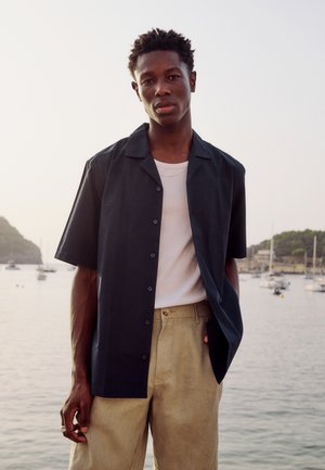 Ung mand står ved en havn iført en kortærmet sort skjorte over en hvid tanktop og beige bukser med både og bakker i baggrunden.