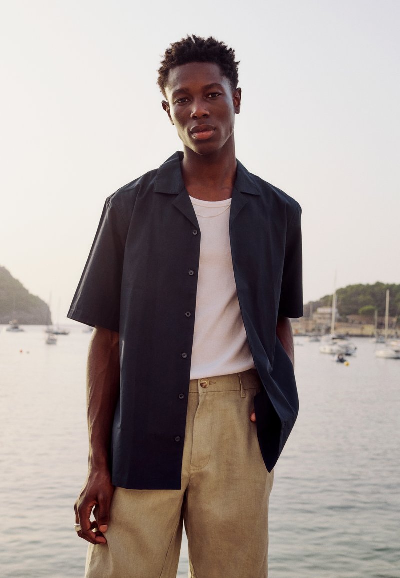 Jeune homme debout près d'un port portant une chemise noire à manches courtes sur un débardeur blanc et un pantalon beige avec des bateaux et des collines en arrière-plan.