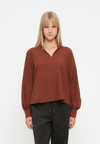 Bruine blouse met lange mouwen, een v-hals, een relaxed fit en verzamelde manchetten. Zachte stof met een gladde textuur, gecombineerd met donkere, gestructureerde broeken.