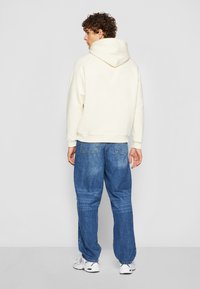 Un sweat à capuche crème avec une poche kangourou, des poignets et un ourlet côtelés. Porté avec un pantalon en denim bleu ample et des chaussures de sport blanches.