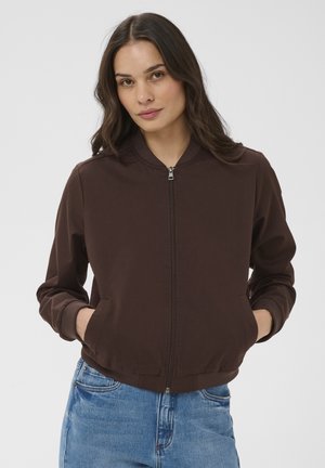 Femeie cu păr lung și întunecat, purtând o jachetă bomber maro închis cu fermoar și blugi albaștri, cu mâinile în buzunare, pe un fundal simplu.