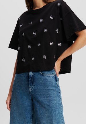 Camiseta de algodón negra de corte crop con un patrón repetitivo de pequeños motivos blancos. Manga corta y un ajuste relajado. Combinada con unos vaqueros de denim de color azul claro.