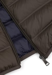 Gros plan sur une veste matelassée marron foncé dézippée montrant une fermeture éclair en métal et une doublure intérieure bleu marine.