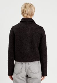 Veste noire texturée, courte, avec un col et des manches longues, portée sur un jean gris clair, vue de dos.