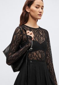 Μαύρο μακρυμάνικο τοπ από δαντέλα πάνω από μαύρο σουτιέν, σετ με μαύρη πλισέ φούστα. Κρατά ένα γυαλιστερό μαύρο τσαντάκι στο χέρι της.