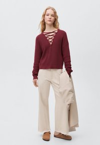 Bordo teksturirana pulover s globokim V-izrezom in križnim vezanjem, v kombinaciji s širokimi bež hlačami z visokim pasom in rjavimi natikači.