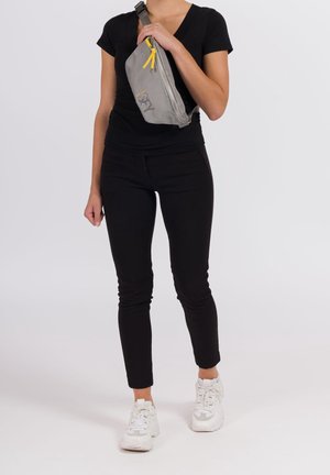 Grå midjeväska med texturerad yta, diagonal form, gul dragkedjehäll och logotyp, bärs över en svart outfit och vita sneakers.