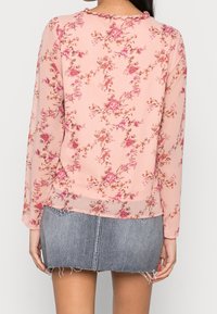 Blusa floreale rosa con maniche lunghe, realizzata in tessuto leggero, con scollatura a scallop e delicati motivi di rose. Indossata sopra una gonna di jeans.