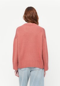 Rosa strikket genser med ribbestrikket kant og mansjetter, som har en avslappet passform. Bæres over lyseblå høytlivsjeans.