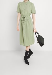 Kobieta w jasnozielonej sukience midi na guziki z paskiem w talii, czarnych botkach na obcasie, trzymająca czarną torebkę na tle białym.