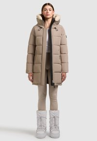 Frau trägt eine beige wattierte Winterjacke mit pelzbesetzter Kapuze, beige Leggings und weiße Winterstiefel, steht vor einem einfarbigen Hintergrund.