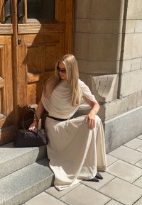 Jasno beżowa bluzka z krótkimi rękawami, drapowana, w zestawieniu z zwiewną beżową spódnicą. Ciemna pleciona torebka spoczywa na kamiennych schodach. Gładkie tekstury, naturalne oświetlenie.