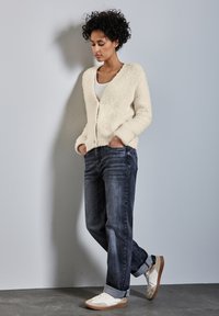 Cremfarbener, flauschiger Cardigan mit V-Ausschnitt und vorderen Knöpfen, kombiniert mit dunkelblauen Jeans und weißen Sneakers mit braunen Akzenten.