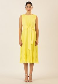 Gelbes plissiertes Kleid mit rundem Halsausschnitt, einem gebundenen Taillengürtel und einem fließenden Saum. Kombiniert mit silbernen Riemchensandalen.