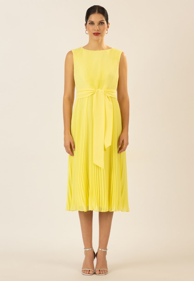 Gelbes plissiertes Kleid mit rundem Halsausschnitt, einem gebundenen Taillengürtel und einem fließenden Saum. Kombiniert mit silbernen Riemchensandalen.