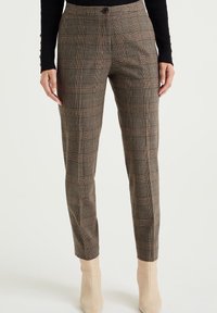 Brown plaid trousers made of textured fabric, featuring a fitted silhouette, button closure, and ankle length. Worn with beige heeled boots.