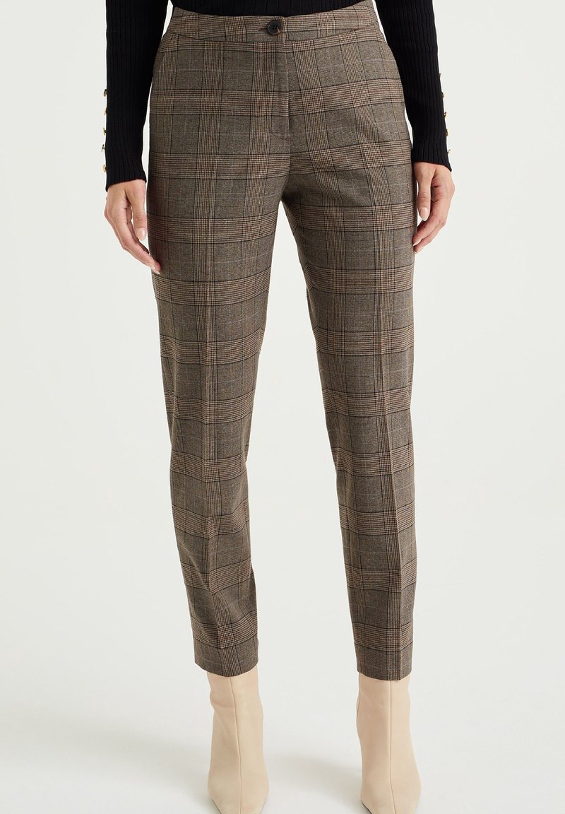 Brown plaid trousers made of textured fabric, featuring a fitted silhouette, button closure, and ankle length. Worn with beige heeled boots.