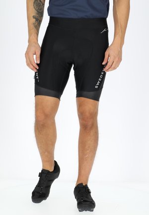 Homme portant un cuissard noir et des chaussures de cyclisme noires, debout devant un fond blanc.