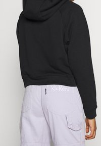 Schwarzer, taillierter Kapuzenpullover, kombiniert mit helllila Shorts, die seitliche Taschen und ein gesticktes "Kickers"-Logo auf der Rückseite haben.