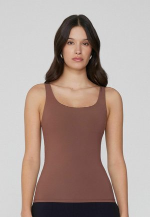 Donna con capelli lunghi e scuri che indossa orecchini a cerchio d'argento e un top marrone aderente senza maniche, in piedi davanti a uno sfondo chiaro e uniforme.