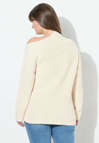 Strickpullover in Creme mit einem schulterfreien Design, geripptem Muster, langen Ärmeln und einem lässigen Schnitt, kombiniert mit hellblauen Jeans.