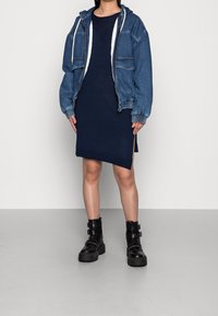 Veste en denim avec capuche portée sur une robe marine avec une fente latérale, associée à des bottines noires à talons épais avec des sangles et des accents.