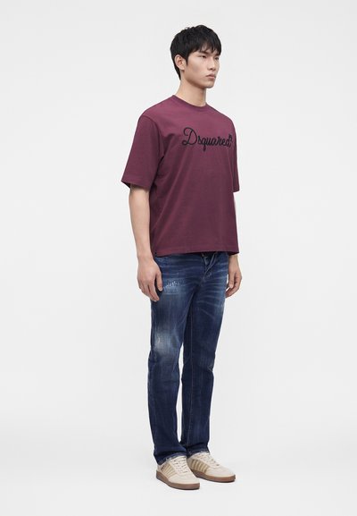 Νέος άνδρας στέκεται φορώντας ένα μπορντό t-shirt "Dsquared2", ξεθωριασμένο μπλε τζιν και μπεζ αθλητικά παπούτσια μπροστά σε απλό λευκό φόντο.