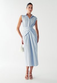 Hele sinine, ilma varruteta midi kleit nupuväärisega kaeluses, kokku kuhjatud vöökoha ja esikottidega. Kandmine koos valgete rihmadega sandaalide ja käekotiga.