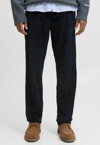 Pantalon en velours côtelé bleu marine avec texture �à côtes verticales, design à jambe droite, porté avec un pull gris et des chaussures de style mocassin marron.