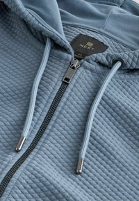 Sudadera con cremallera de color azul claro con un patrón geométrico texturizado, que presenta una etiqueta de logo negra, cremallera plateada y cordones a juego.