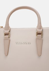 Sac à main beige avec une finition texturée, double anses supérieures, quincaillerie dorée et nom de marque en relief sur le devant.